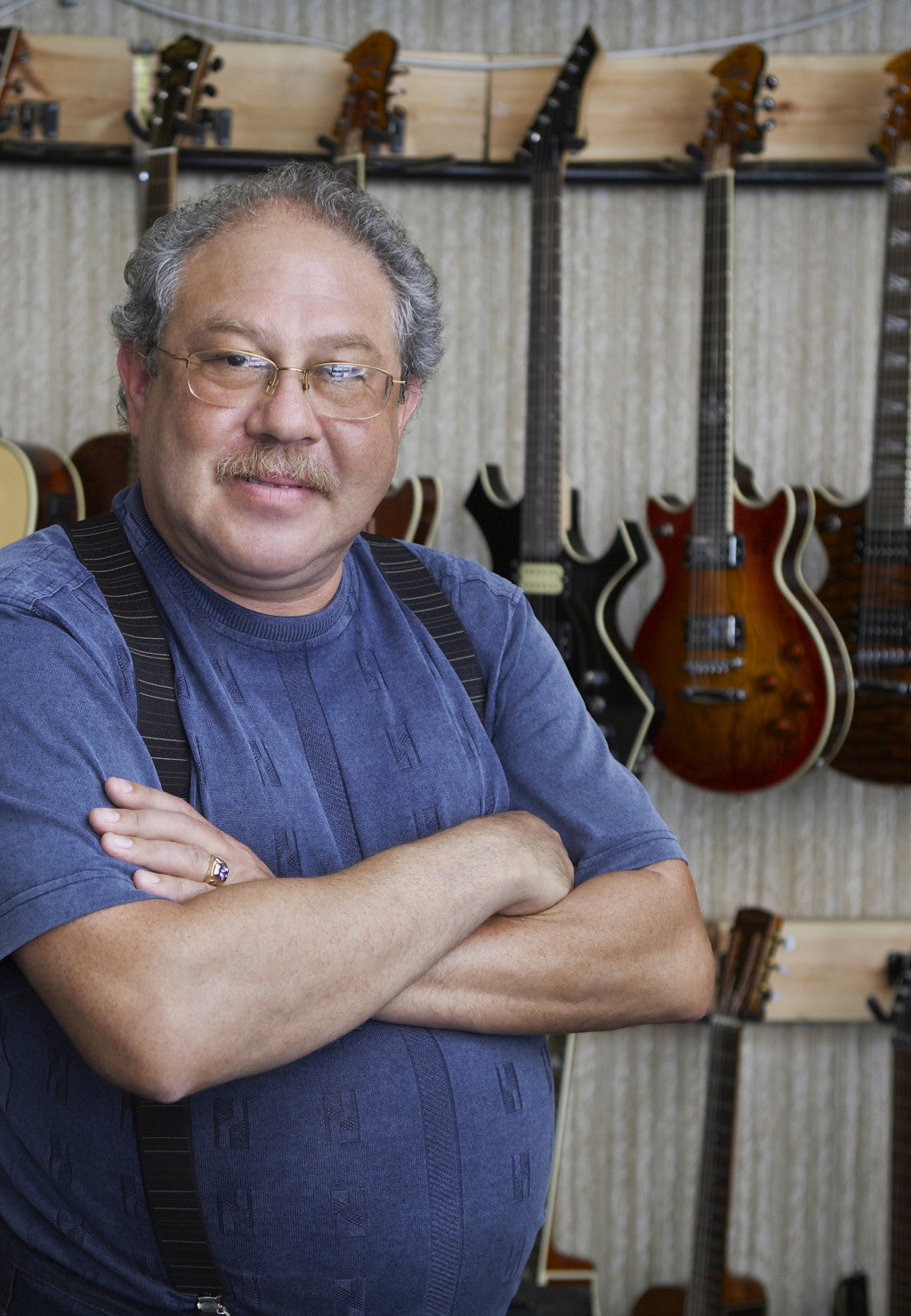 A man with gray hair, glasses, and a mustache stands with arms crossed, smiling. He wears a blue shirt and suspenders. Behind him, various electric guitars are displayed on a wall-mounted rack.