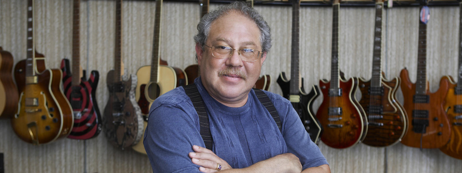 A man with gray hair, glasses, and a mustache stands with arms crossed in front of a wall displaying various electric and acoustic guitars.